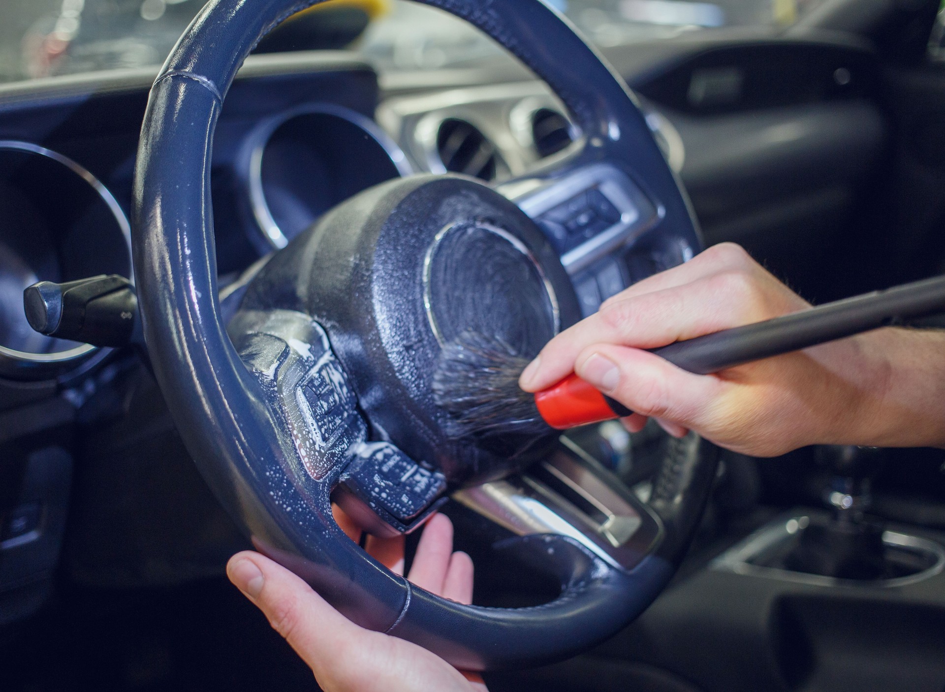 Car detailing and the concept of car interior care. A young man cleans the steering wheel of a modern car using a special brush. Dry cleaning of the car interior, close-up, selective focus.